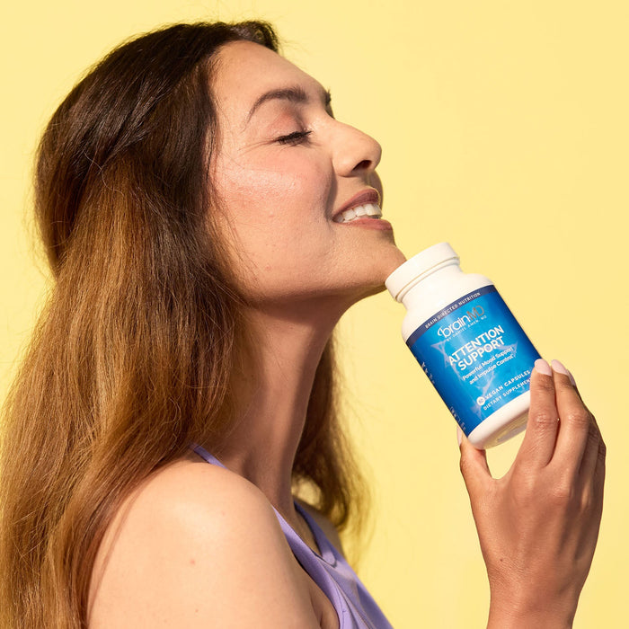 BrainMD Attention Support: woman in front of a yellow background holds a supplement bottle against her chin