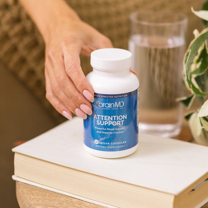 BrainMD Attention Support: a woman's hand holds a supplement bottle on a book with a blurred interior background