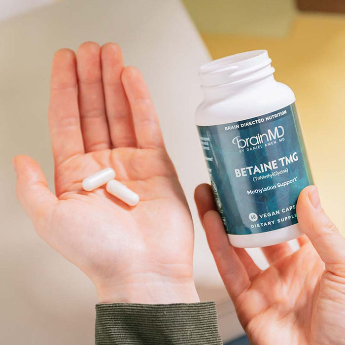 BrainMD Betaine TMG: person holding a blue supplement bottle in one hand and white capsules in the other