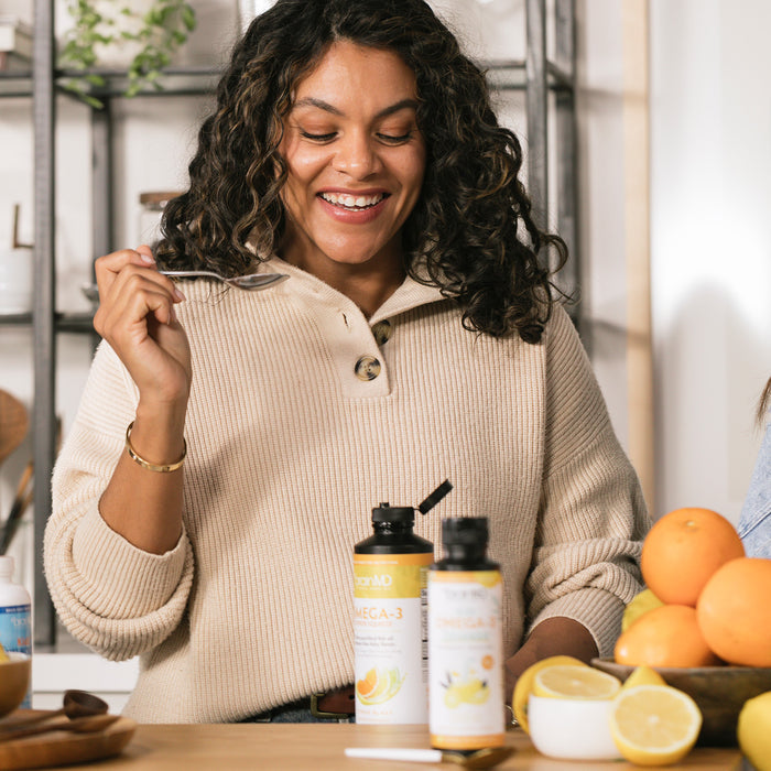 BrainMD Daily Essentials with Squeeze Bundle: smiling woman holds spoon in kitchen, supplement bottles next to sliced oranges