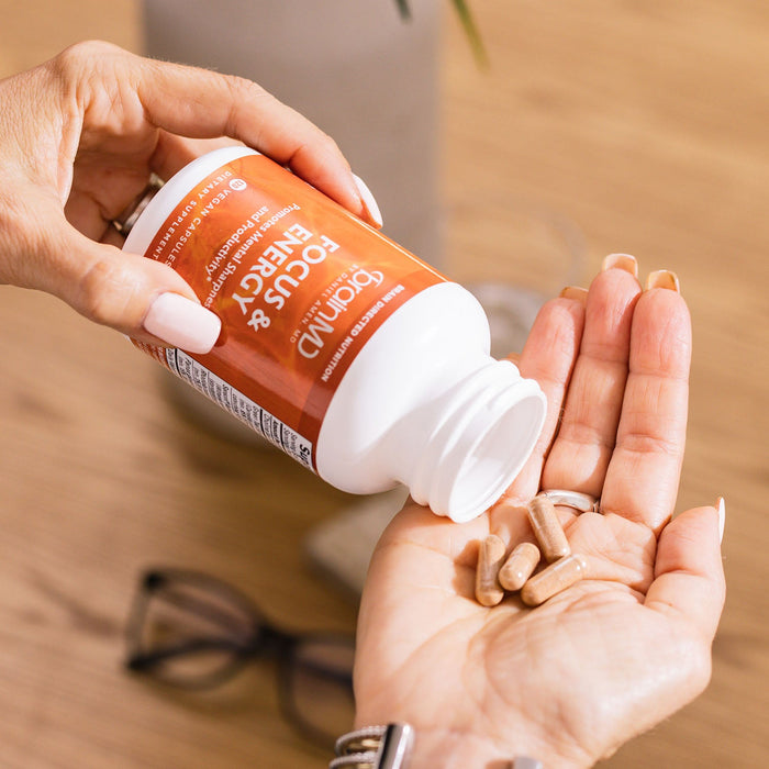 BrainMD Focus & Energy: woman pours capsules into her palm from an orange supplement bottle against a blurred background