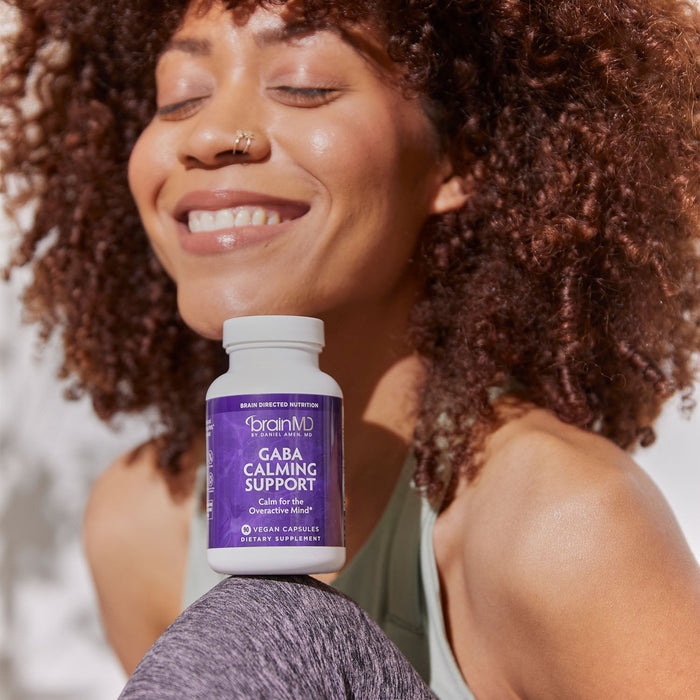 BrainMD GABA Calming Support: smiling woman balances purple supplement bottle between her chin and knee