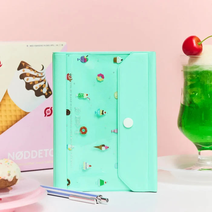 The closed cover against pink with an ice-cream poster, a green cream soda and a pen