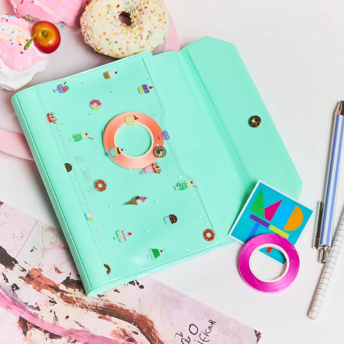 The cover on pink with a doughnut, a roll of tape and a pen beside it