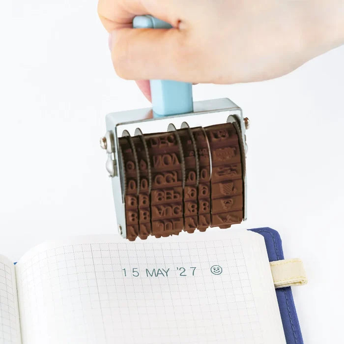 A hand pressing the stamp onto a grid page, leaving 15 MAY '27 behind