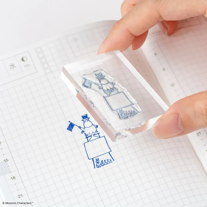 Hands stamping the mounted design onto a grid page, the blue impression showing below