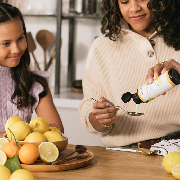 BrainMD Kids' Omega-3 Squeeze: woman in kitchen pouring liquid from a supplement bottle into a spoon as daughter looks on