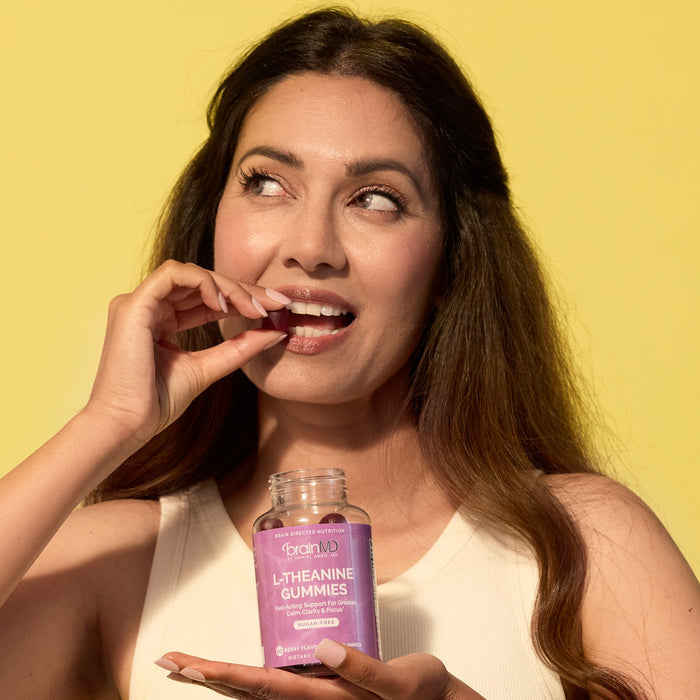 BrainMD L-Theanine Gummies: woman chewing a gummy and holding a purple supplement bottle against a yellow background