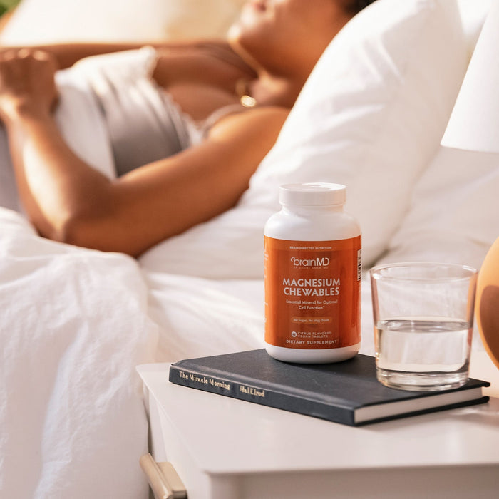 BrainMD Magnesium Chewables: blurred woman sleeps in the background, orange supplement bottle and water glass in foreground