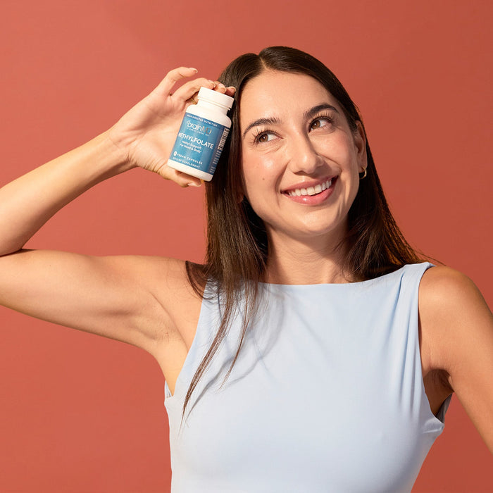 BrainMD Methylfolate: woman holding supplement bottle to her forehead, standing in front of an orange background