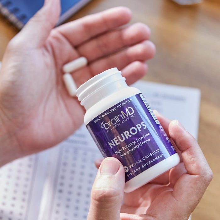 BrainMD Neuro-PS: person pouring capsules into hand from a purple supplement bottle, blurred desk background