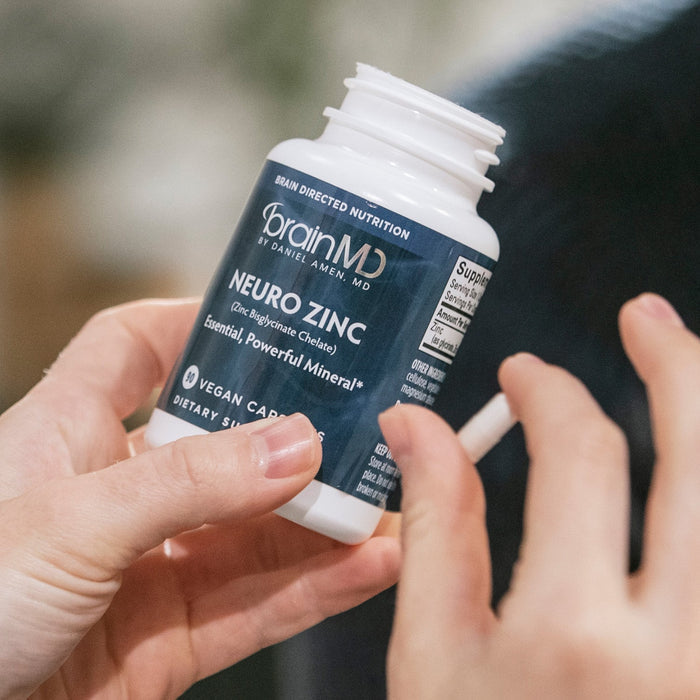 BrainMD Neuro Zinc: person holding gray supplement bottle and white capsule against a blurred interior background