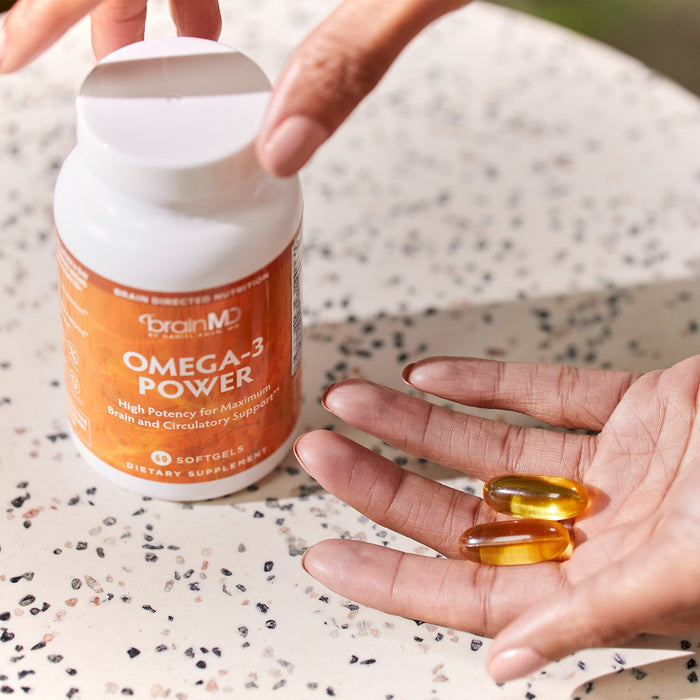 BrainMD Omega-3 Power: orange supplement bottle with person holding softgels in their hand, speckled countertop background