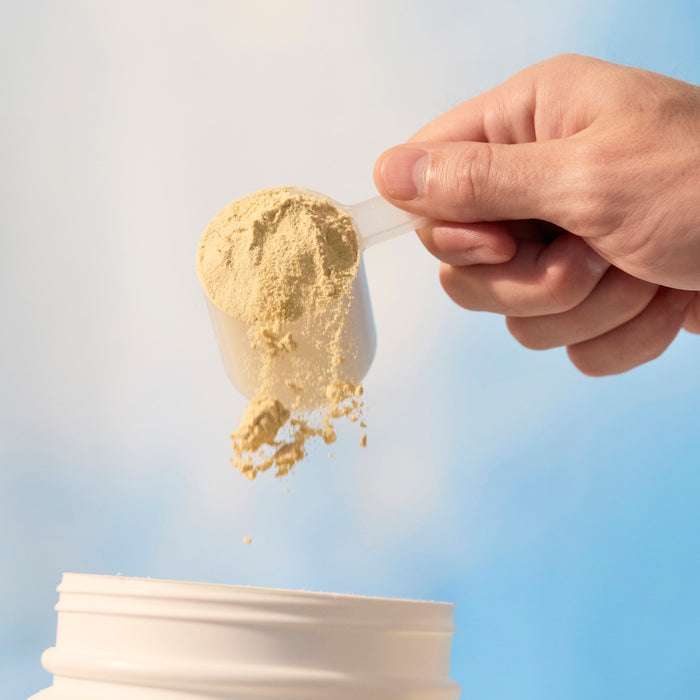 Hand pouring scoop of protein powder into a container against a sky background.