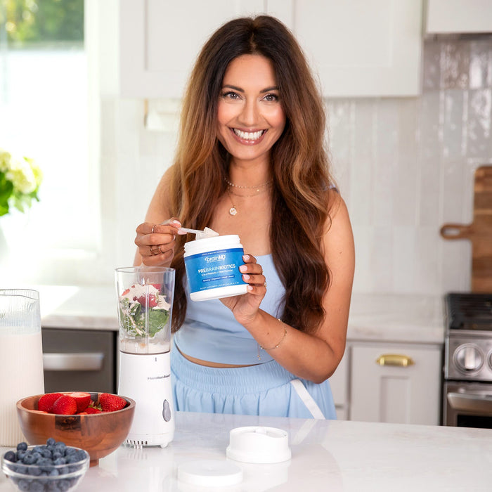 BrainMD PreBrainBiotics: smiling woman scooping some powder from a blue supplement container into a blender, interior kitchen
