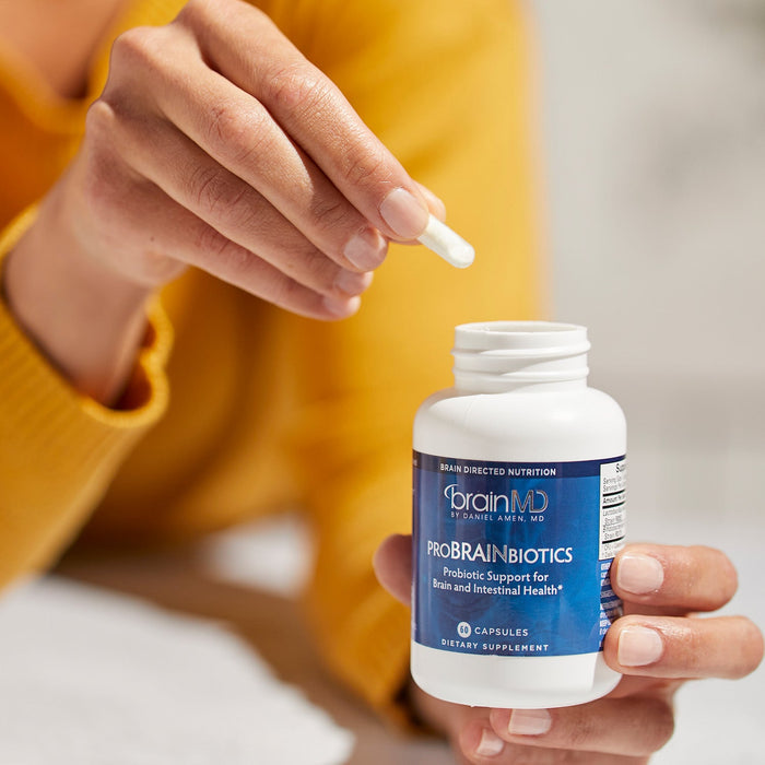BrainMD ProBrainBiotics: woman with white capsule in one hand and a blue supplement bottle in the other, blurred background