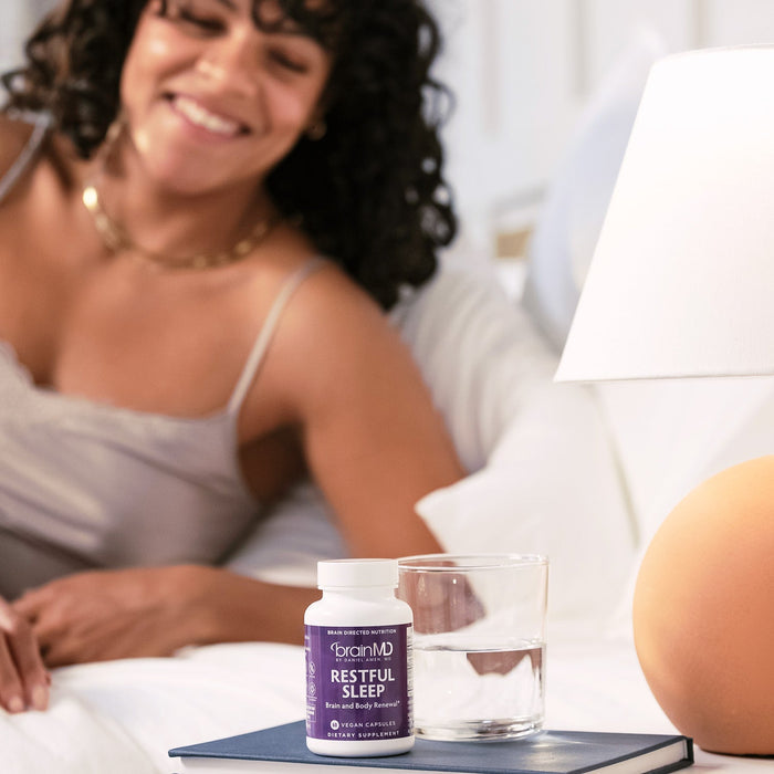 BrainMD Restful Sleep: blurry image of a smiling woman in background, purple supplement bottle on nightstand in foreground