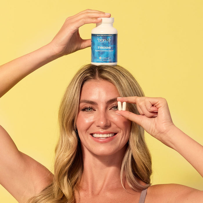 BrainMD Tyrosine: woman holding blue supplement bottle on her head & two capsules in front of her eye, yellow background