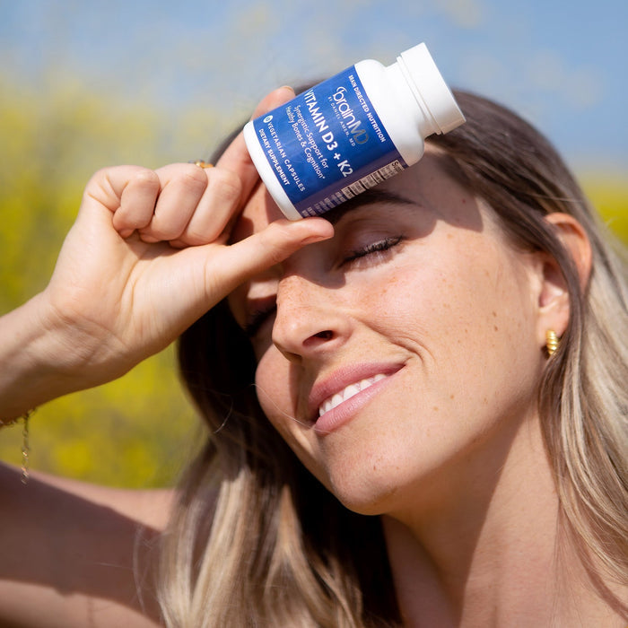 BrainMD Vitamin D3 + K2: woman holds blue supplement bottle to forehead against yellow exterior background