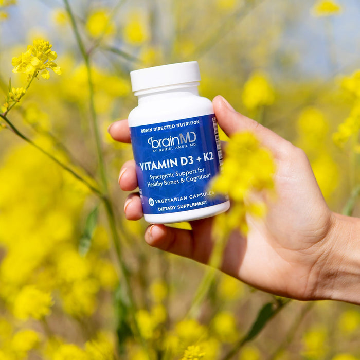 BrainMD Vitamin D3 + K2: woman holds blue supplement bottle against a blurred yellow exterior background
